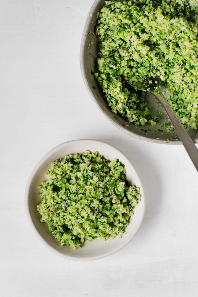 A small bowl of broccoli tabbouleh is accompanied by a skillet filled with the same dish. They rest on a white surface.