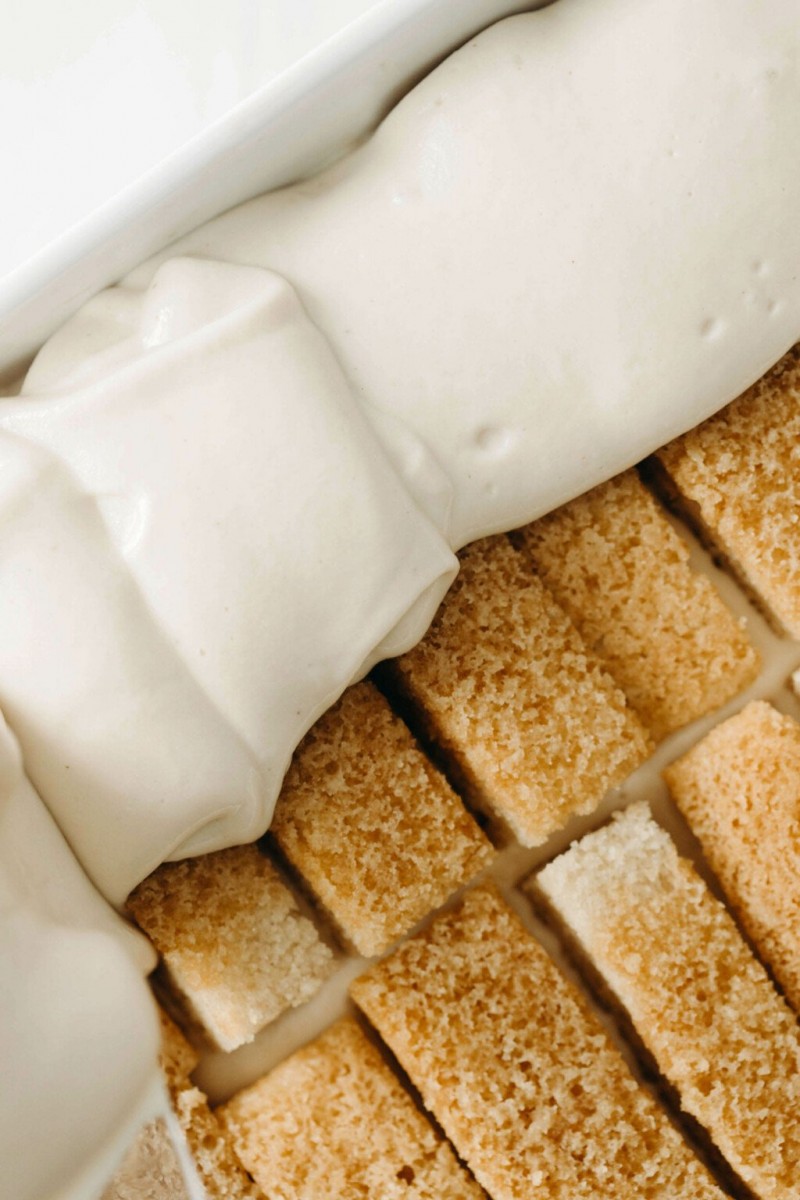 Rectangular slices of vanilla cake are being topped with a layer of a rich, creamy white mixture.