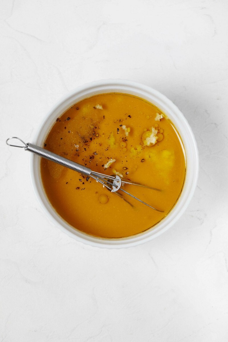 An overhead image of a small white bowl, which is filled with a pale orange vinaigrette, garlic, salt, and pepper.