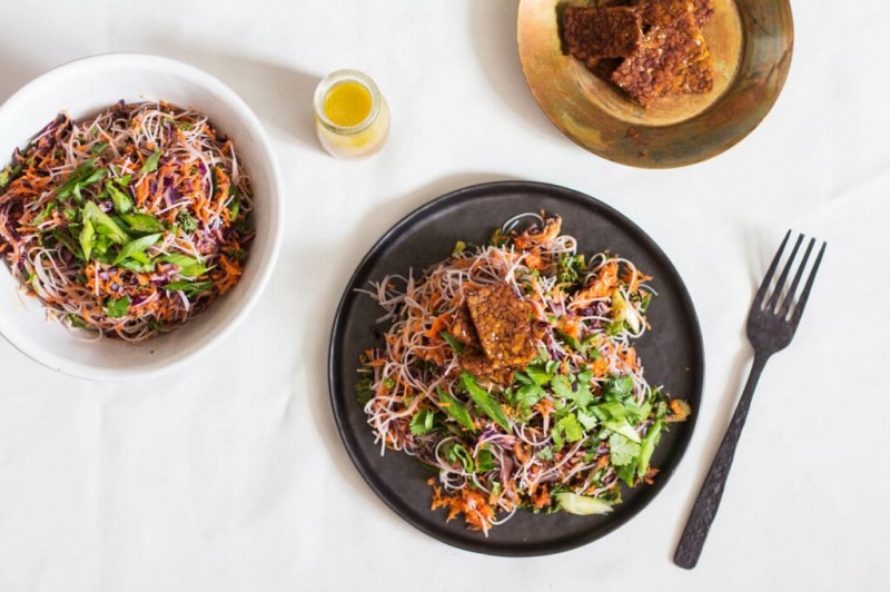 A vegan kelp noodle cabbage salad has been plated on a round, black matte plate. A small bowl of seared tempeh pieces rests to the top right.