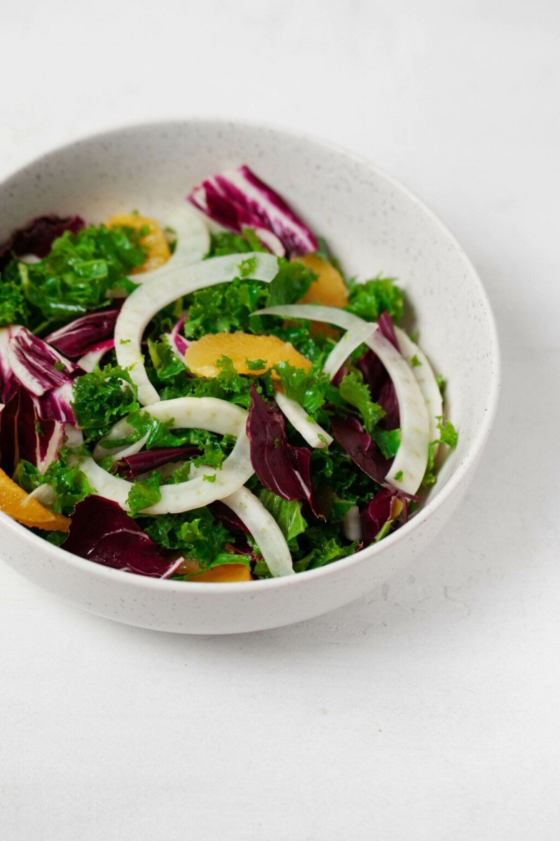 An overhead image of a large white mixing bowl, which is filled with shaved fennel, kale, radicchio, and orange sections.