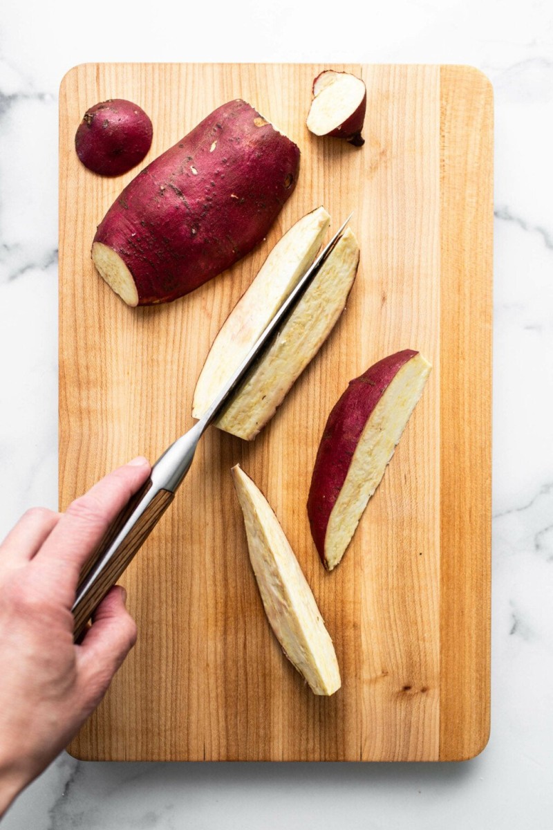 An overhead image of whole and sliced Japanese sweet potatoes on a light brown cutting board.